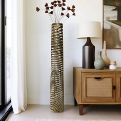 a tall metal vase sitting on top of a table next to a lamp and window