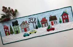 a quilted wall hanging with houses and cars on it's side, along with pine branches