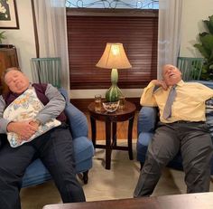 two men are sleeping on blue chairs in the living room, one is holding a pillow