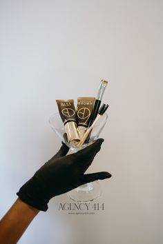 a person in black gloves holding up a glass vase with some makeup products on it