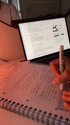 a person is holding a pen and writing on a notebook with a laptop in the background