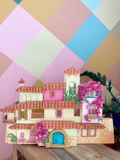 a model of a house with pink flowers on the roof and windows, in front of a multi - colored wall