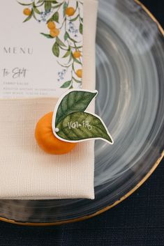 an orange sitting on top of a glass plate next to a napkin and place card