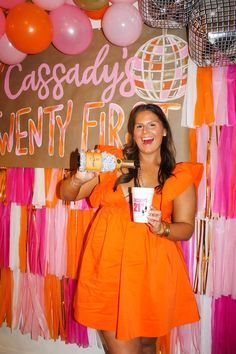 a woman in an orange dress is holding a cup and posing for the camera with balloons behind her