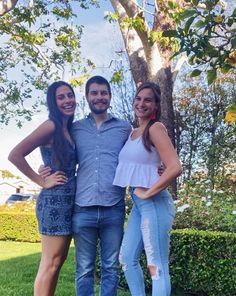 two women and a man standing next to each other in front of a tree on the grass