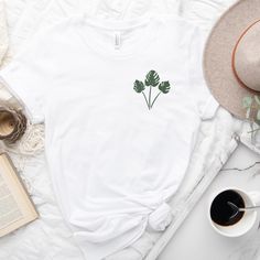 a white t - shirt with a green plant on it next to a cup of coffee and an open book