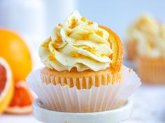 a cupcake with white frosting and orange zest on top sits in front of some grapefruits