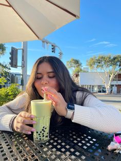 a woman sitting at an outdoor table drinking from a tall glass with a straw in her mouth