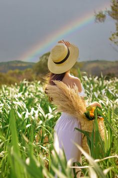 a woman in a white dress and straw hat standing in tall grass with a rainbow in the background