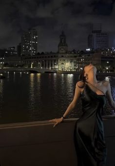 a woman standing in front of a body of water at night with buildings in the background