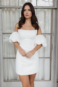 a woman standing in front of a window with her hands on her hips wearing a white dress