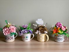 three coffee mugs with flowers in them sitting on a wooden table next to a wall