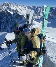 two people standing on top of a snow covered slope holding skis and poles with mountains in the background