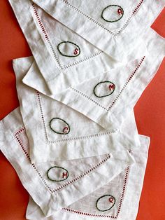 six white napkins with embroidered designs on them sitting on a red tableclothed surface