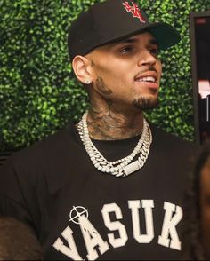 a man wearing a black shirt and hat standing in front of a wall with plants