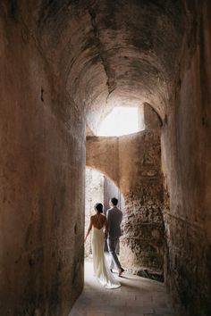 a man and woman are walking through an alley way in the stone building with light coming from behind them