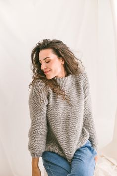 a woman sitting on top of a bed next to a white wall and wearing a gray sweater