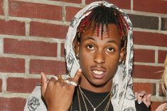 a young man with dreadlocks standing in front of a brick wall and pointing to his finger