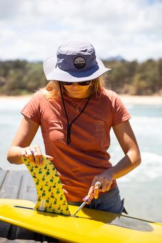 a woman sitting on top of a yellow surfboard next to the ocean holding a pair of scissors