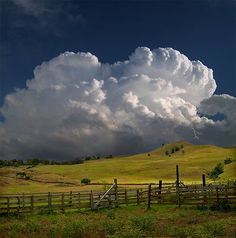 a large cloud is in the sky over a farm field with a wooden fence and green grass