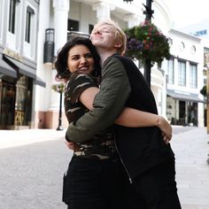 a man and woman hugging on the street in front of some buildings with flowers behind them