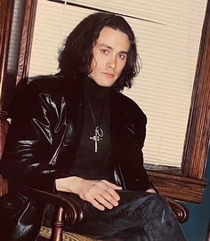 a man sitting in a chair wearing a black shirt and leather jacket with long hair