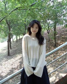 a woman is standing on the steps in front of some trees and bushes, wearing a white sweater