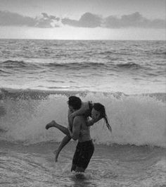 a man carrying a woman into the ocean while standing on top of a surfboard