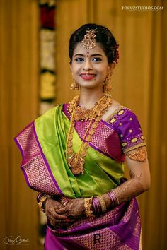 a woman in a purple and green sari with gold jewelry on her neck, smiling at the camera