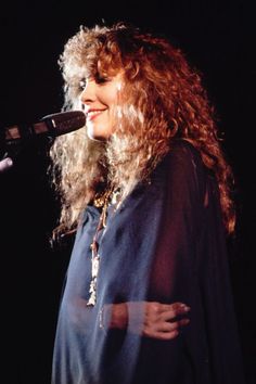 a woman with long curly hair standing in front of a microphone and holding her arms around her chest