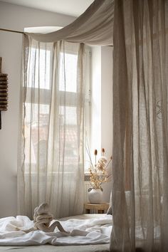 an unmade bed with sheer curtains and flowers in the window sill next to it