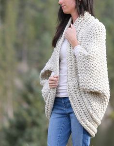 a woman is walking in the woods wearing a white cardigan sweater and blue jeans