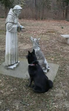 a black dog sitting next to a statue in the grass with a cat looking at it