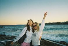 two young women standing next to each other near the ocean with their arms in the air