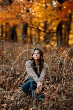 a woman sitting on the ground in front of trees with autumn leaves around her and looking at the camera