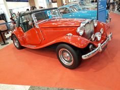 an old red car is on display in a building with people looking at the cars