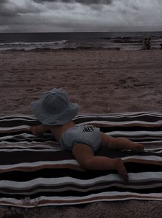 a baby laying on top of a blanket next to the ocean under a cloudy sky