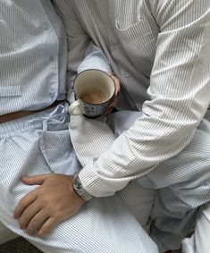 a man in pajamas holding a cup of coffee while sitting on a bench with his arm wrapped around another person's chest