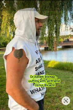 a man wearing a white hoodie standing in front of a lake with trees and grass