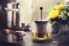 a glass tea pot with a black handle on a counter next to a vase filled with yellow flowers