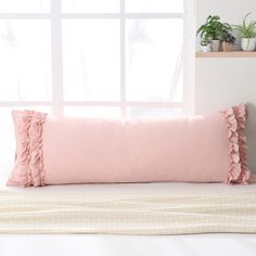 a pink pillow sitting on top of a white bed next to a window with potted plants