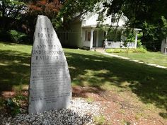 a large stone marker in front of a white house with trees and grass around it