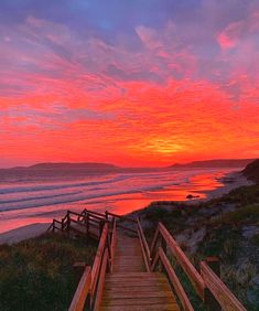 a wooden walkway leading to the beach at sunset with clouds in the sky and water behind it