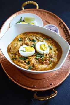 a bowl of soup with boiled eggs and garnishes on top, sitting on a copper platter