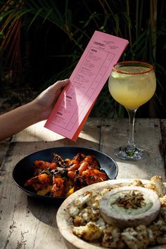 a person holding up a menu next to a plate of food and a glass of wine