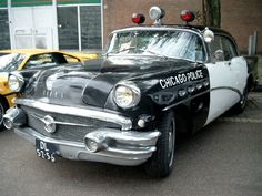 an old black and white police car parked next to a yellow taxi cab in front of a brick building