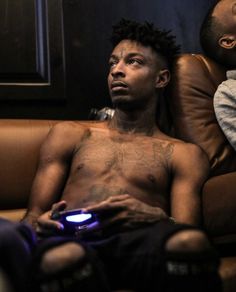 a shirtless man sitting on a couch holding a game controller in his hand and looking at the camera