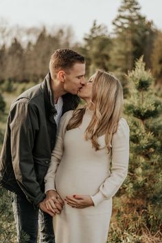 a pregnant couple kissing while standing next to each other in front of some pine trees