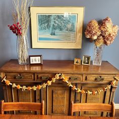 a wooden table topped with a vase filled with flowers next to a painting and two framed pictures