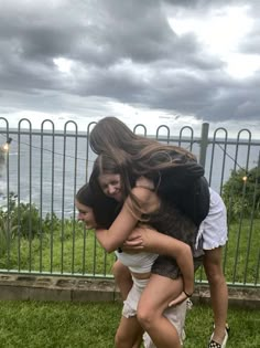 two girls are hugging each other in front of a fence and the ocean behind them